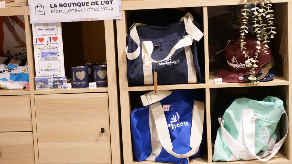 Wooden display shelf with Montgenèvre souvenirs: blue and white tote bags, burgundy cap, mugs and cards reading 'I ♥ Montgenèvre', and travel postcards.