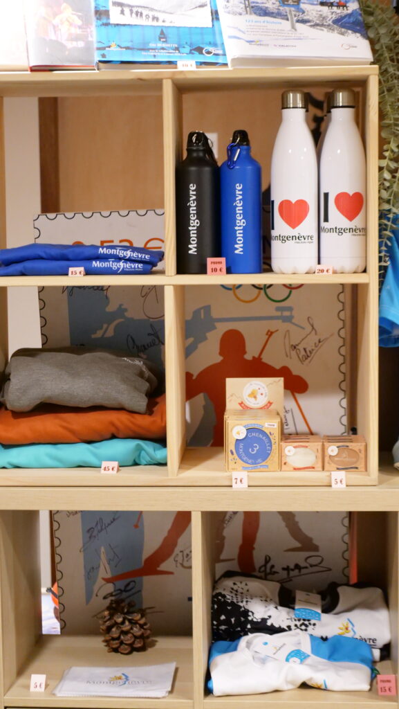 Store display shelf with Montgenèvre-branded water bottles, apparel, and souvenirs on wooden cubbies behind glass panels at a shop.