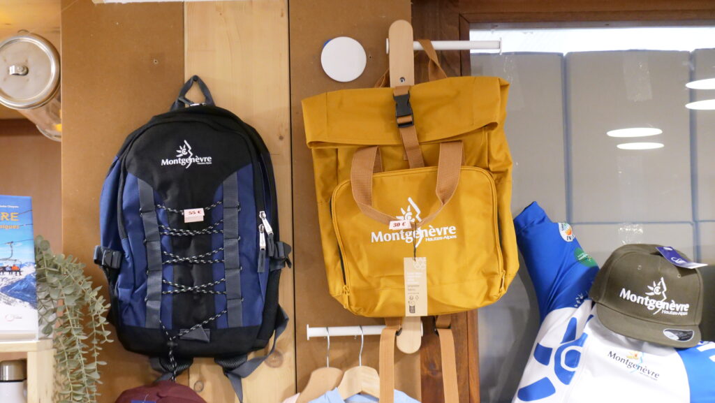 Blue and black Montgenèvre hiking backpack and mustard yellow Montgenèvre bag hanging on a wooden display shelf in a shop.] ,