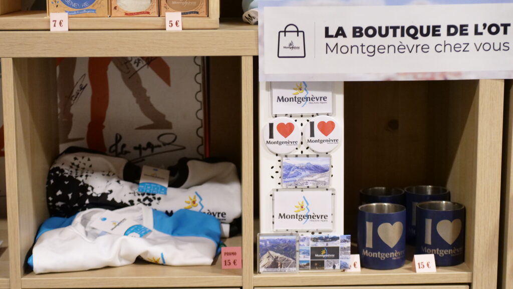 Store shelf with Montgenèvre souvenirs: socks, magnets, postcards, and mugs featuring 'I ❤️ Montgenèvre' logos and a large OT sign above.